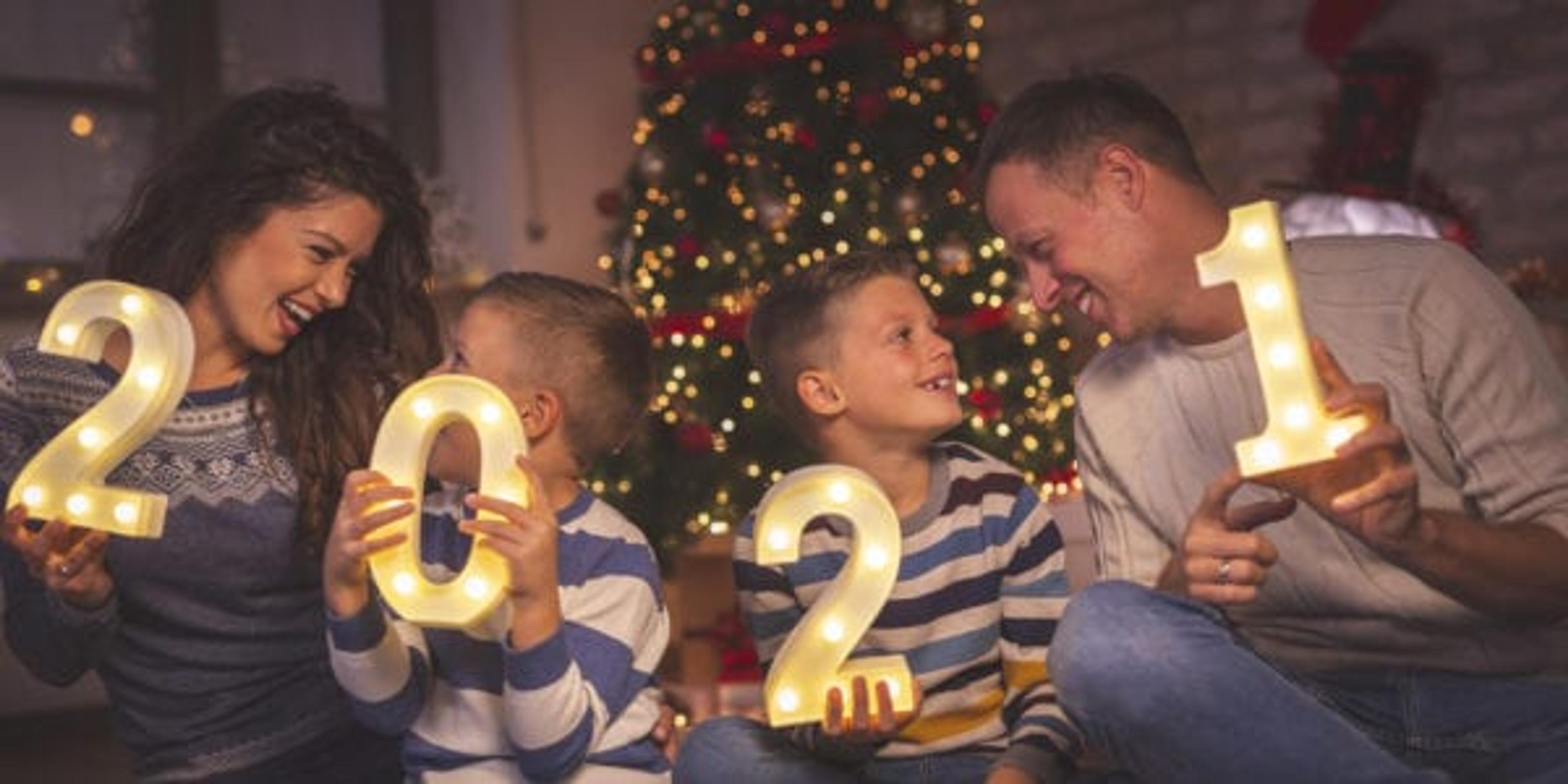 Parents celebrating New Years Eve at home with kids, sitting by the Christmas tree, holding illuminative numbers 2021.
